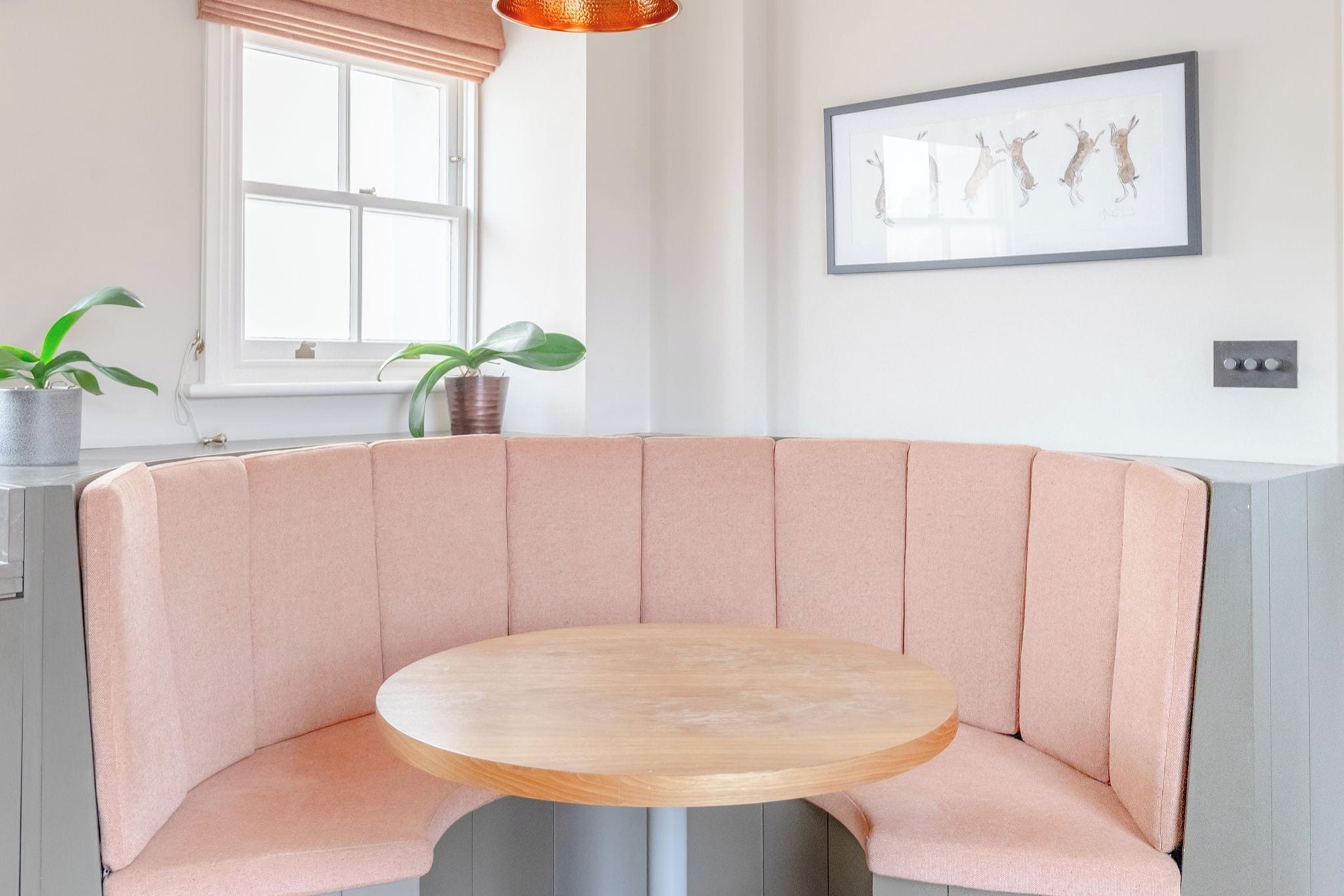 Un coin salon avec une table ronde, une fenêtre avec des stores et un tableau encadré au mur.
