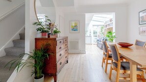 A modern dining area with a wooden table and chairs, a staircase with a plant, and a view of a library. - Beautifully Designed 2BD House - by Queen's Park! (London)
