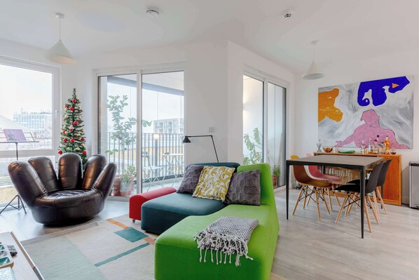 A modern living room with a green sofa, a leather chair, a dining area with a table and chairs, a Christmas tree, and large windows. - Bright 1 BDR Flat Balcony Heart of Brixton (Brixton)