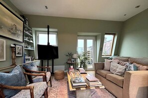 Un salon comprenant un canapé, des fauteuils, une table basse avec des livres, un téléviseur et une bibliothèque.