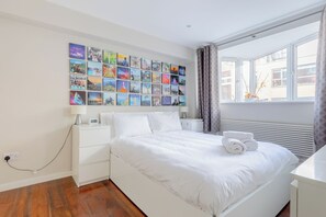A hotel room with a large bed, a bedside table, a wall of colorful pictures, a window with curtains, and a power outlet. - Chic High-End 1BD Flat - Near Borough Market! (London)