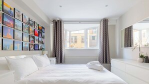 Quarto de hotel com uma cama grande, uma parede de quadros, uma janela com cortinas, um radiador e uma mesa de cabeceira com um espelho.