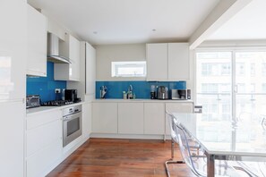 A modern kitchen with white cabinets, a blue backsplash, and a view of the city through a large window. - Chic High-End 1BD Flat - Near Borough Market! (London)