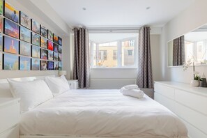 A hotel room with a large bed, a wall of framed photos, a window with curtains, and a bedside table with a mirror.