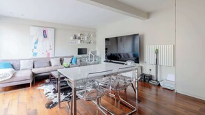 Uma sala de estar moderna com uma mesa de jantar de vidro, cadeiras transparentes, um sofá, uma TV de tela plana e um radiador.