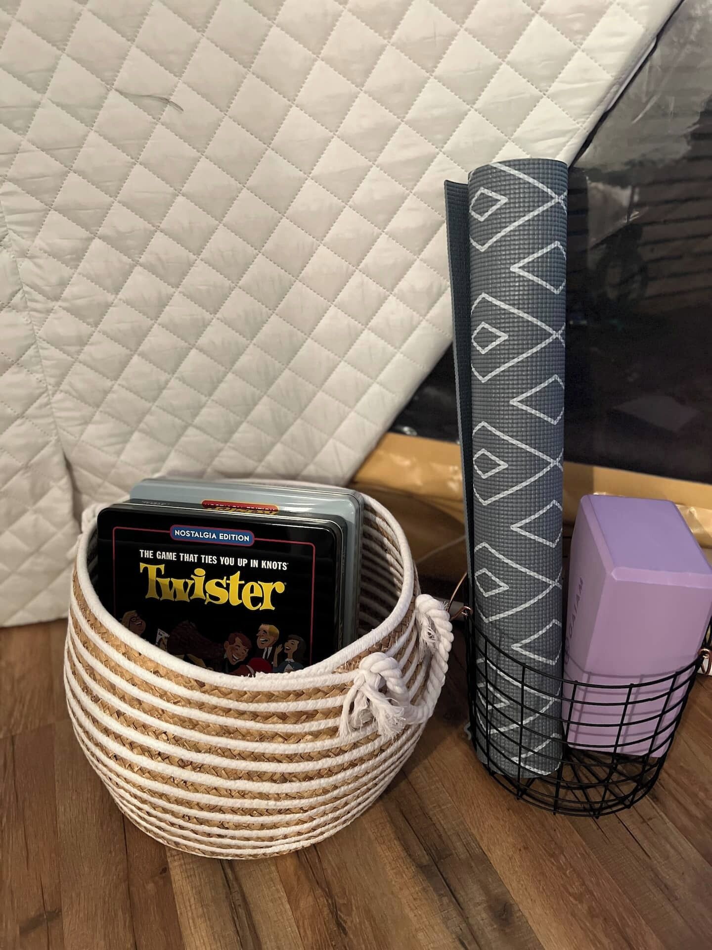 A woven basket containing a board game titled "Twister" and a black and gray striped item, possibly a board game or accessory, next to a purple object in a wire basket.