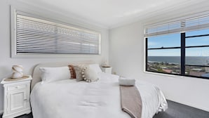 A bedroom with a large bed, white walls, a view of the ocean, and a window with blinds.