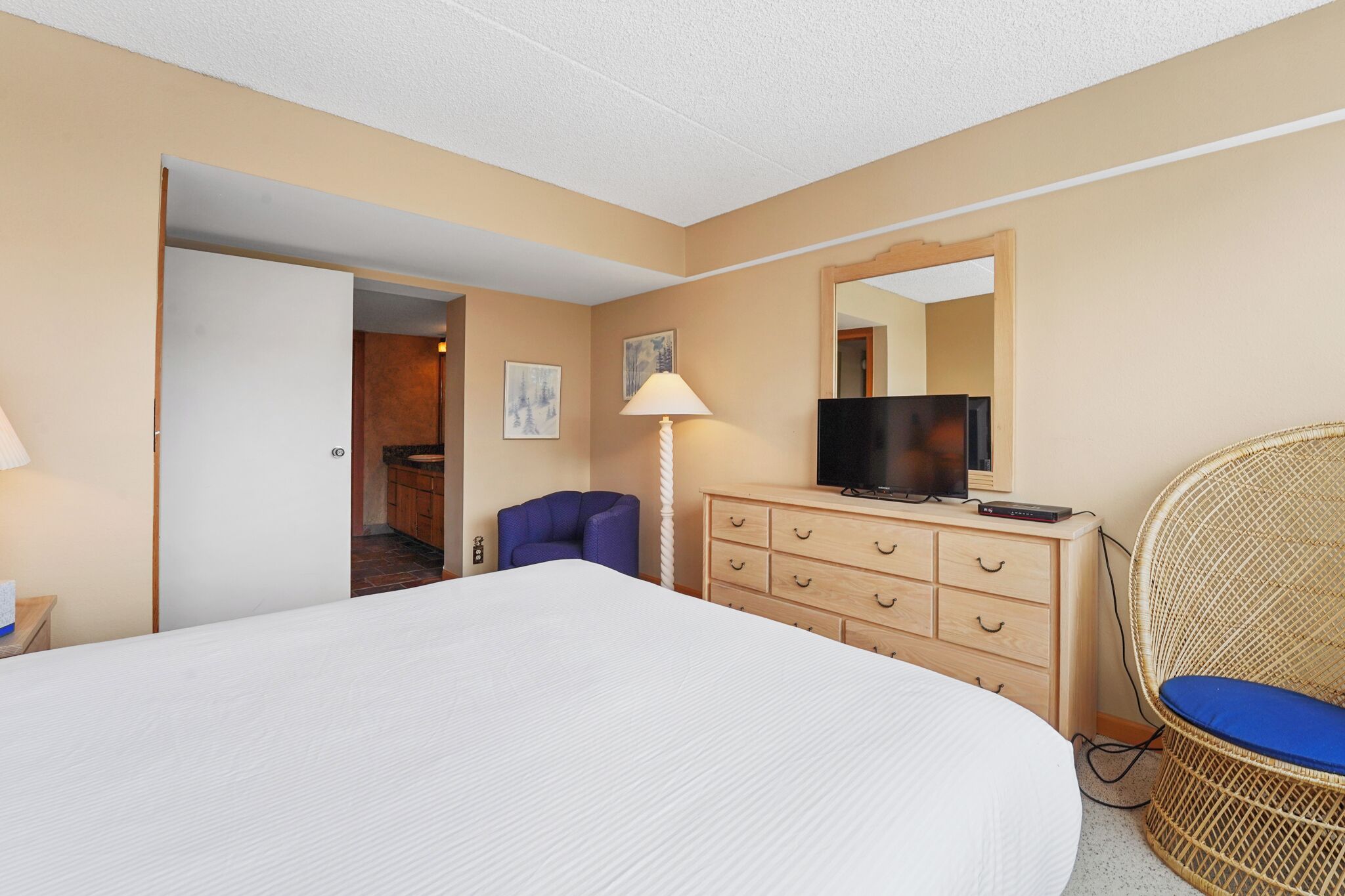 Habitación de hotel con una cama grande, un televisor en una cómoda de madera, una silla de mimbre con un cojín azul, y una puerta que da a un baño.