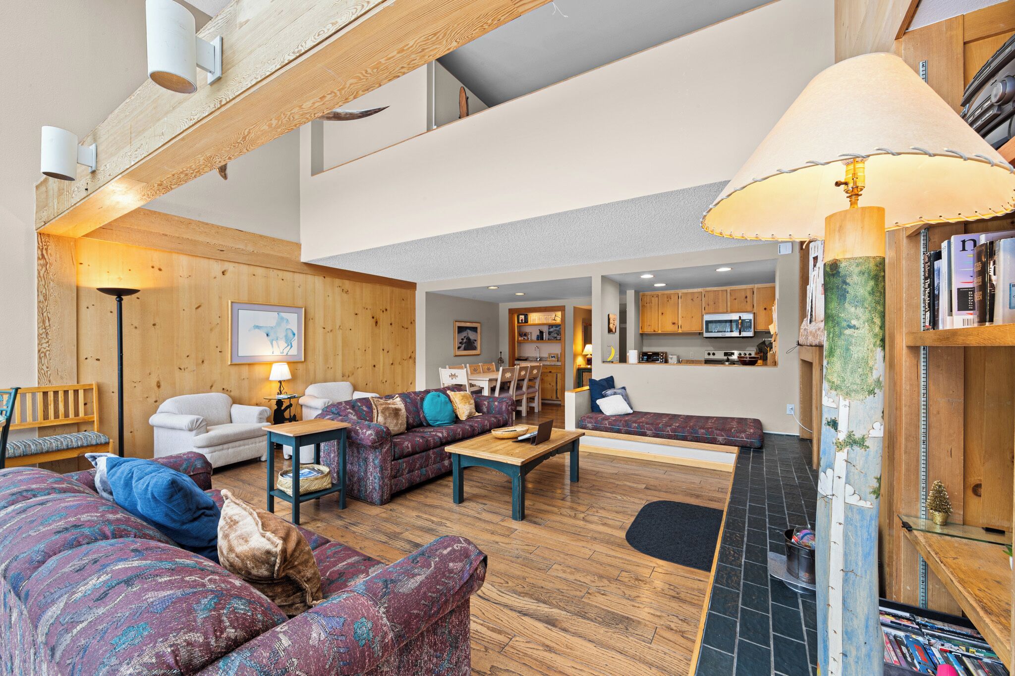 A spacious living area with a high ceiling, wooden beams, and a mix of seating including sofas and a chair. A kitchen is visible in the background.
