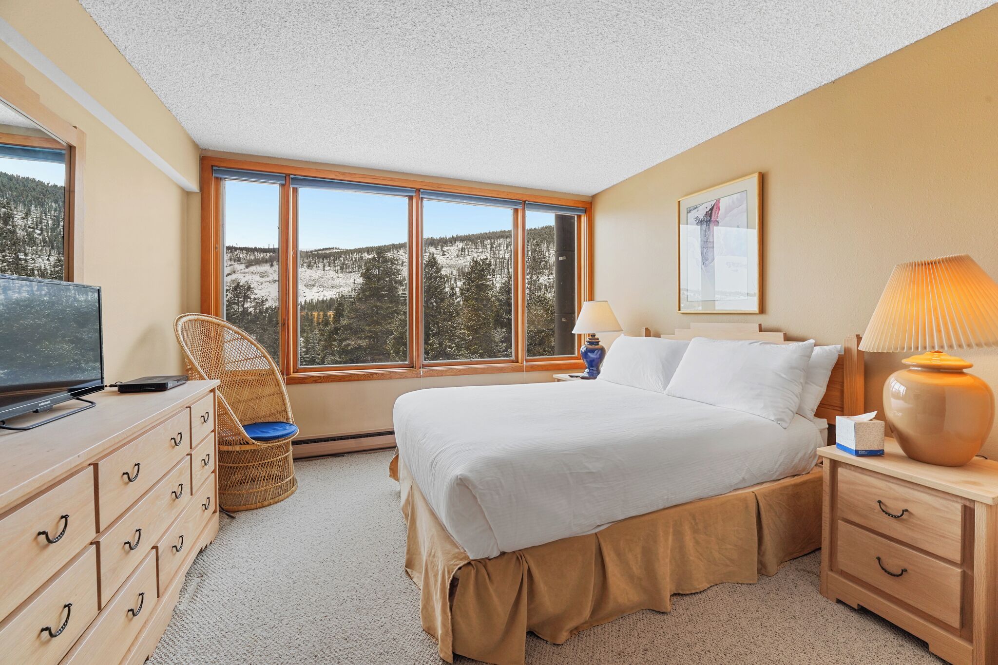Une chambre d’hôtel avec une grande fenêtre, un lit, une commode, une chaise et une lampe.