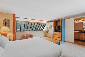 A bedroom with a large bed, a desk, a chair, a window with a view of trees, and a kitchenette with cabinets and a stove.