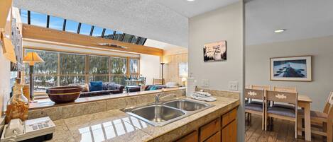 A modern kitchen with granite countertops, stainless steel sinks, and wooden cabinets. There is a dining area with a table and chairs, and a large window offering a view of trees.