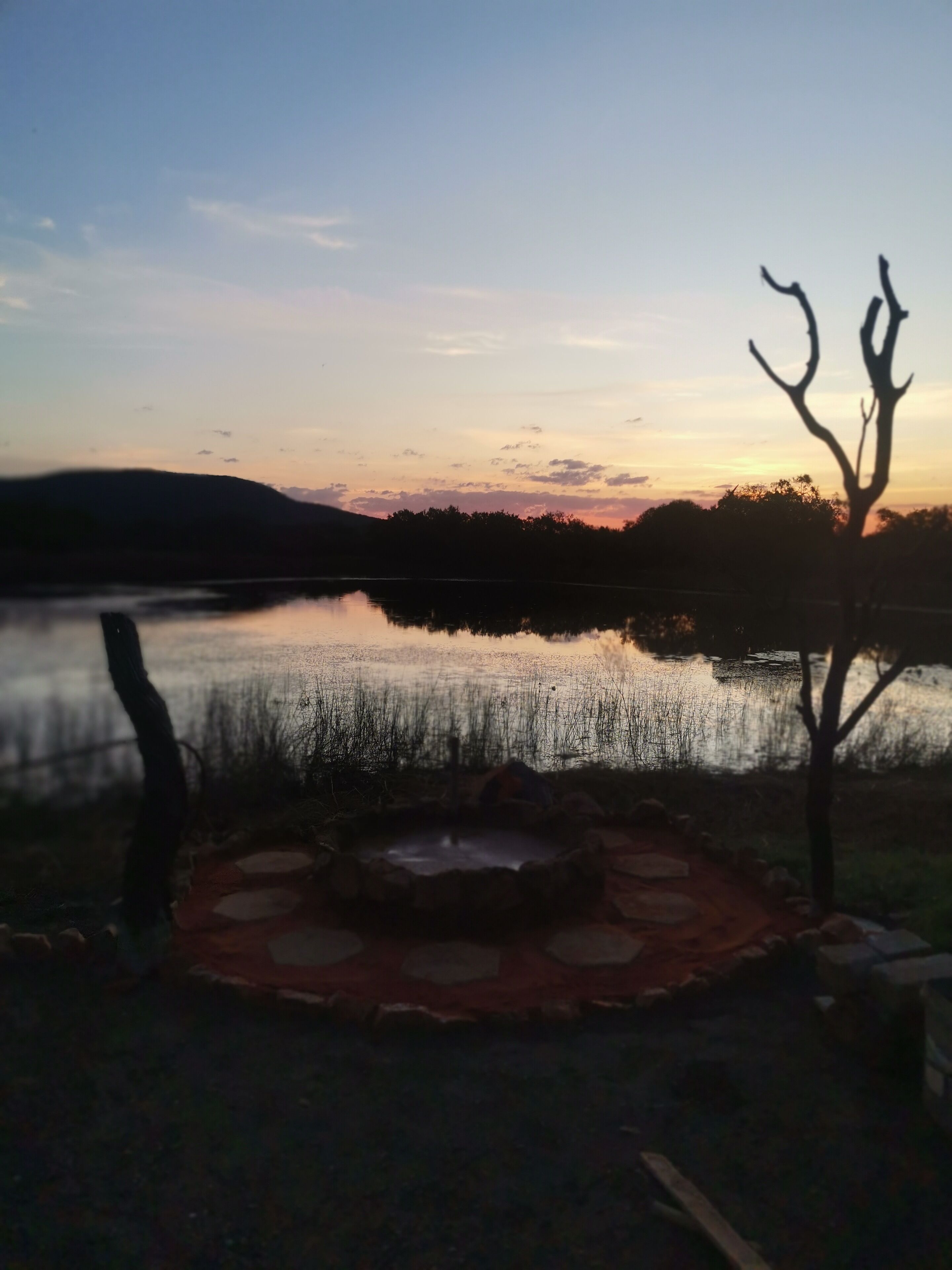Cenário à beira do lago, com uma lareira rústica em primeiro plano e a silhueta de uma árvore contra um céu ao pôr do sol.