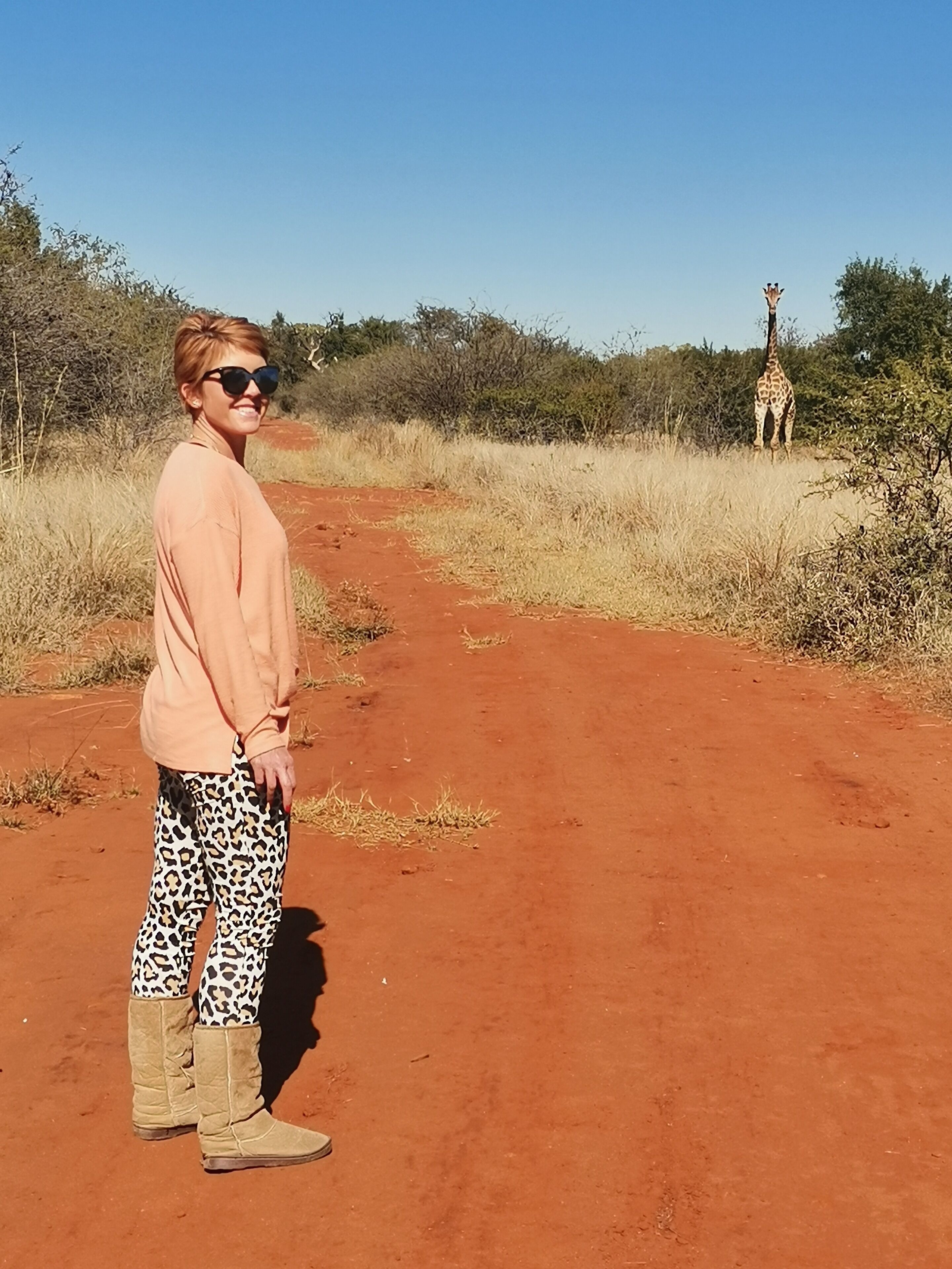 A person standing on a dirt path with a giraffe in the background.