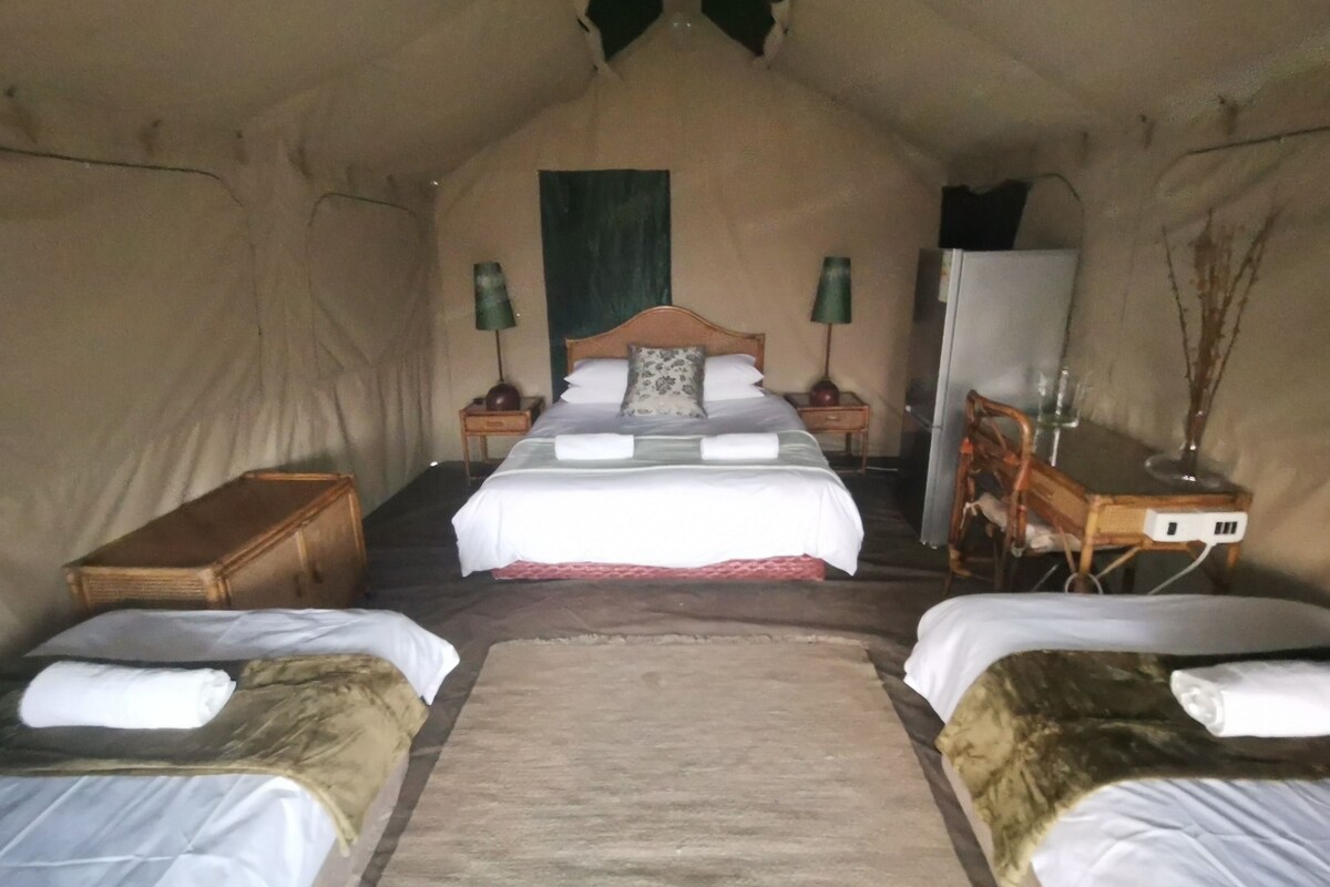 A cozy interior of a tent with a central bed, two smaller beds on either side, a wooden nightstand with a lamp, and a small table with a vase.
