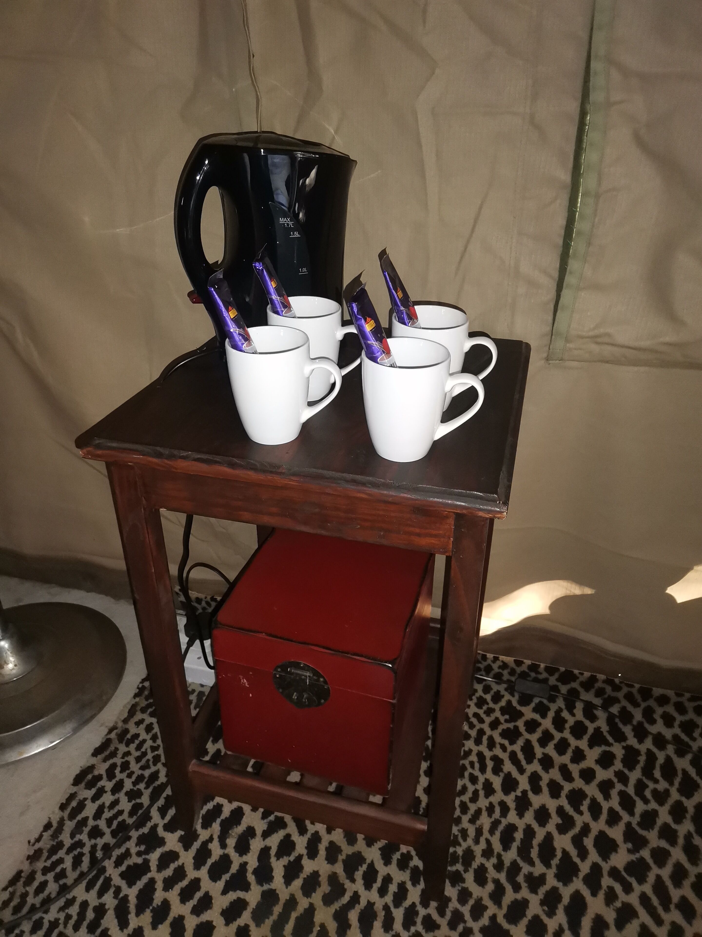 Una mesa con tazas blancas, una cafetera negra, una caja roja y un piso estampado de leopardo.