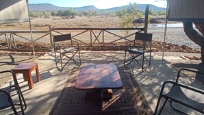 A tented area with a table and chairs, a patterned rug, and a view of a dry landscape with hills and sparse vegetation.