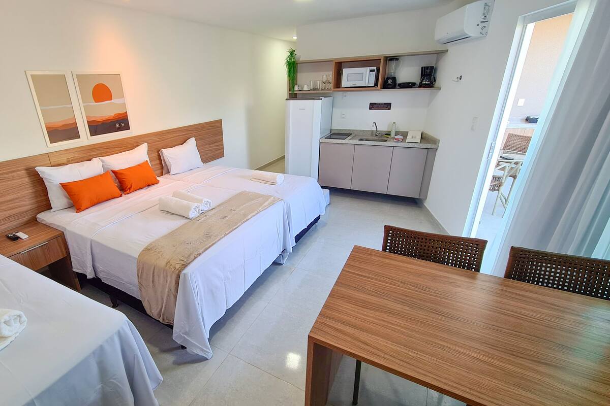 Habitación de hotel con cama doble, cama individual, mesa de madera, kitchenette y obras de arte en la pared.