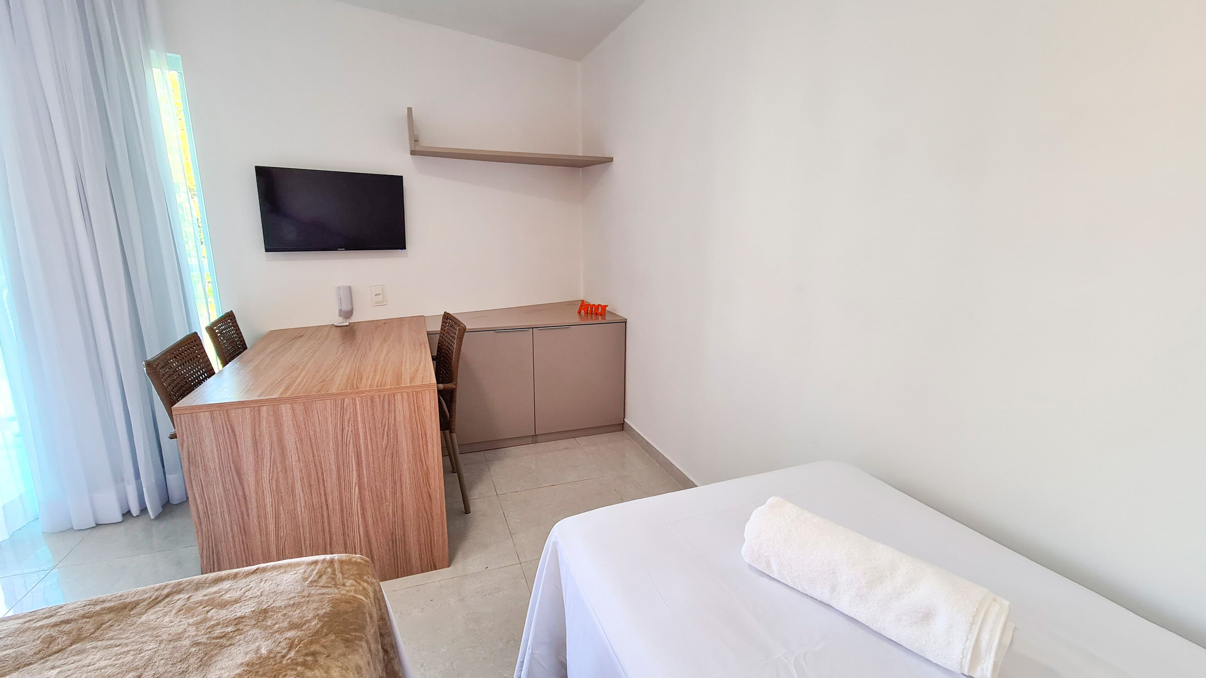 A hotel room with a wooden desk, a television mounted on the wall, a small shelf, a bed with a white blanket, and a window with curtains.