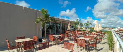 An outdoor patio with wicker tables and chairs, a palm tree, and a view of the city skyline.