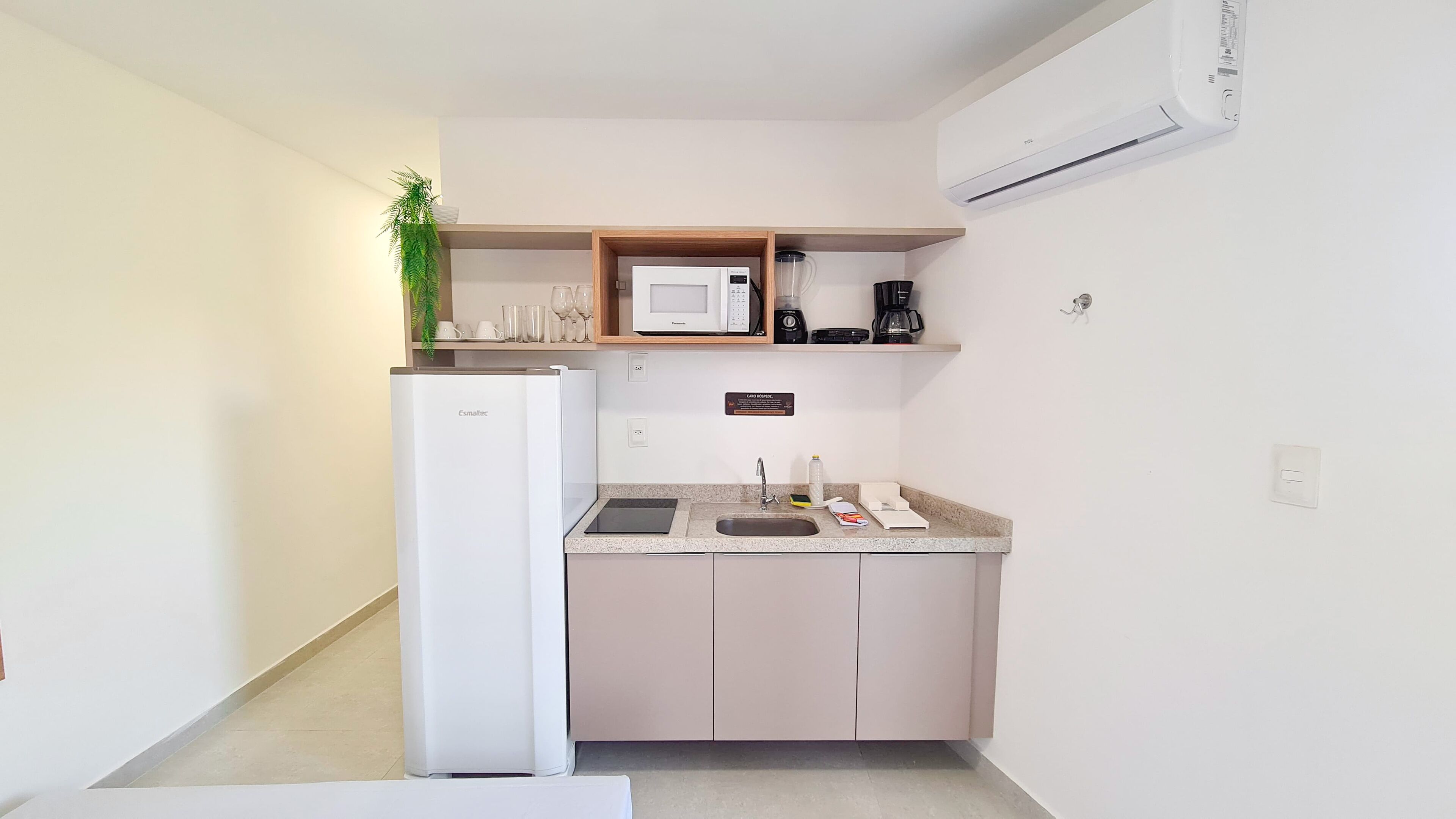 A compact kitchen with a refrigerator, microwave, coffee maker, and a small shelf with cups and a plant.