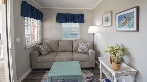 Un salon avec un canapé beige, une table basse turquoise et une console blanche ornée d’une plante. Il y a deux fenêtres avec des rideaux bleus, un lampadaire et des tableaux encadrés accrochés au mur.