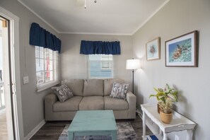 Un salon avec un canapé beige, une table basse turquoise et une console blanche ornée d’une plante. Il y a deux fenêtres avec des rideaux bleus, un lampadaire et des tableaux encadrés accrochés au mur.
