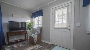 Un salon avec un téléviseur posé sur une console en bois, un fauteuil avec une couverture, une porte avec une fenêtre et un tapis bleu.