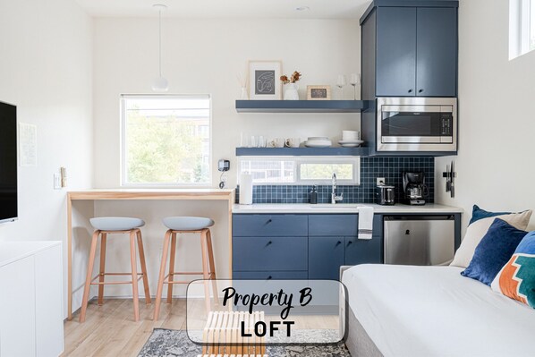 A modern loft bedroom with a large bed, a desk with two stools, a kitchenette with blue cabinets, and a view of greenery outside.