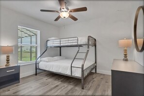 A bunk bed with a ceiling fan, a bedside table with a lamp, and a window with blinds.