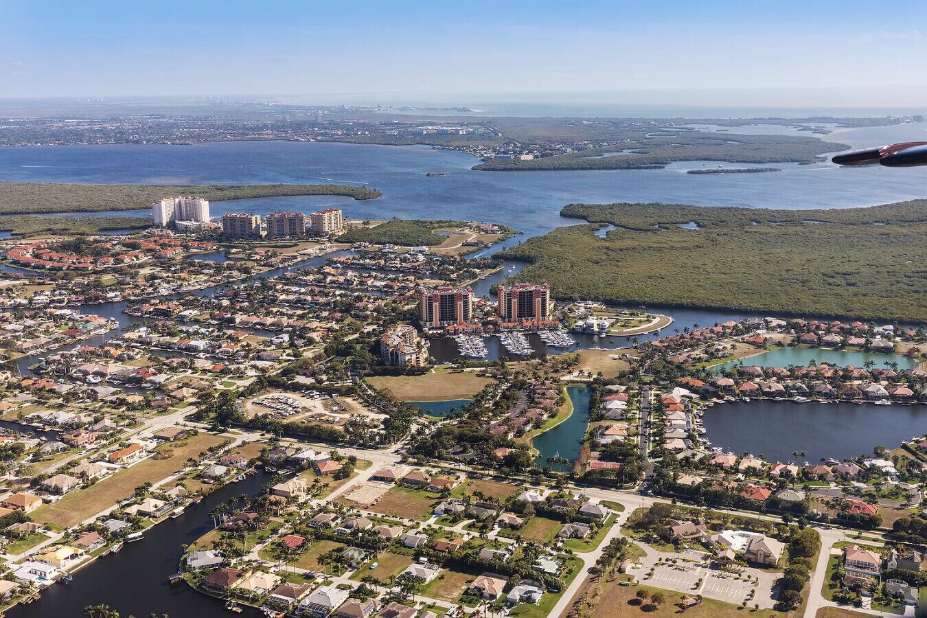An aerial view of a coastal residential area with multiple buildings, including high-rise condos and smaller houses, surrounded by water bodies and greenery.