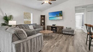 A living room with a sectional sofa, a coffee table, a TV, and a ceiling fan. - New Build W/ Heated Pool! Perfect for 2 families! (Cape Coral)