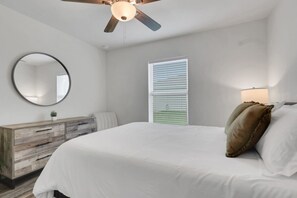 A bedroom with a bed, a wooden dresser, a round mirror, a ceiling fan, and a window with blinds.