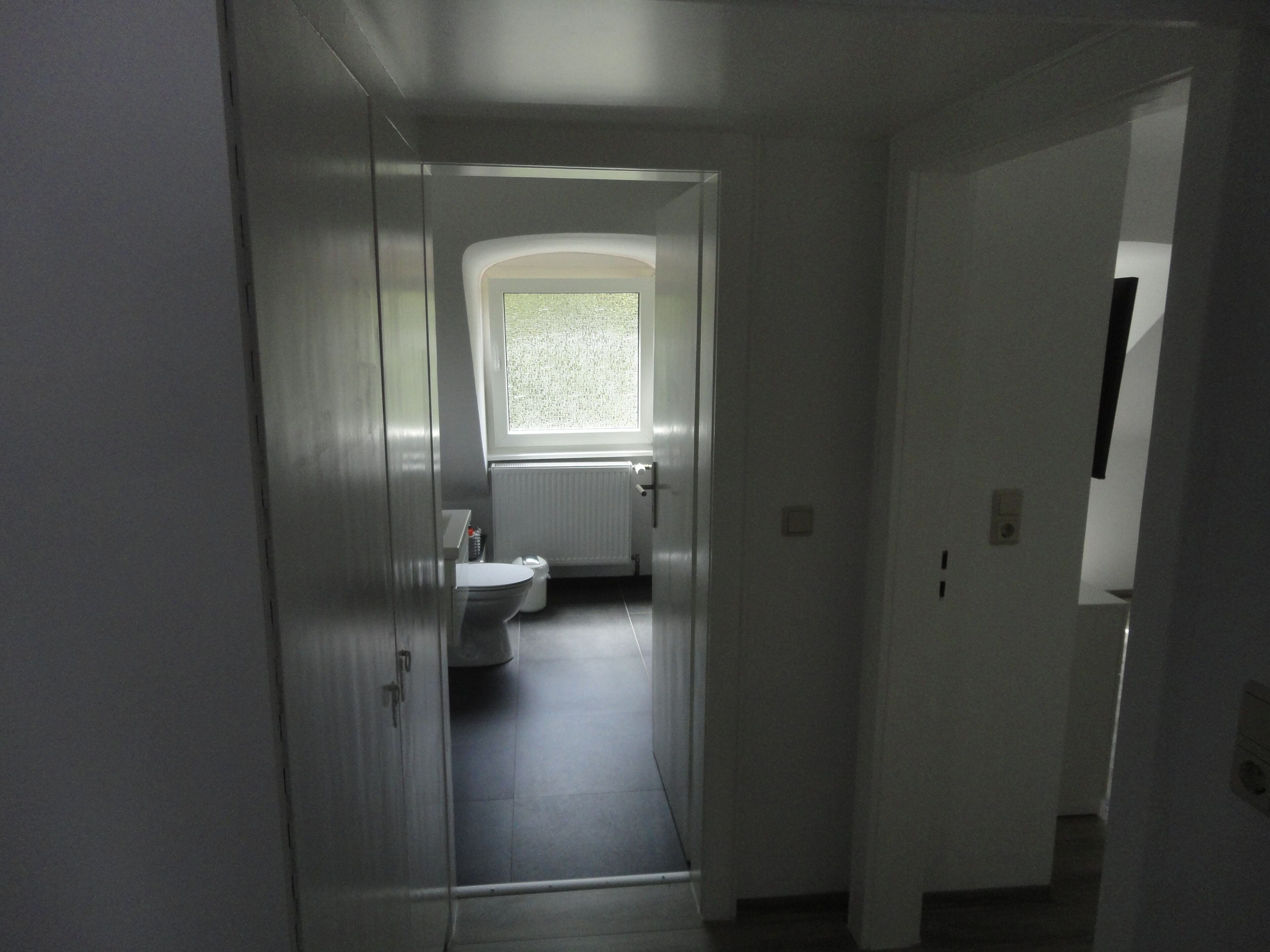Een smalle gang die leidt naar een badkamer met een toilet, een radiator en een raam.