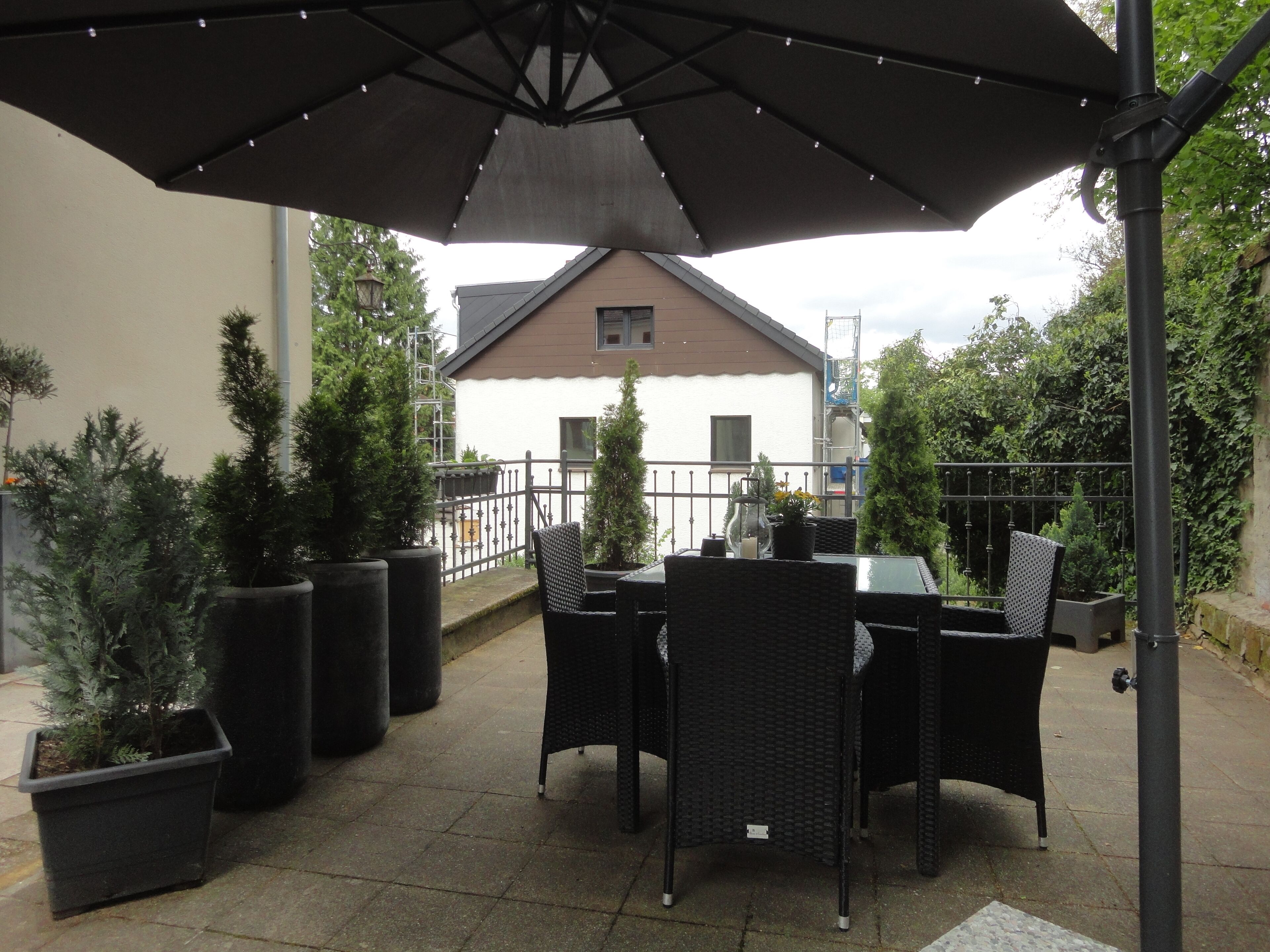 Eine Terrasse im Freien mit Tisch und Stühlen, umgeben von Blumentöpfen und einem großen Sonnenschirm.