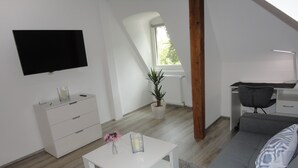 Ein modernes Wohnzimmer mit einem weißen Sofa, einem weißen Couchtisch, einem an der Wand montierten Fernseher, einem Schreibtisch mit Stuhl und einer Topfpflanze.