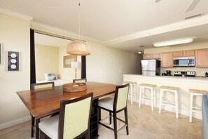 A dining area with a wooden table and chairs, a woven light fixture, and a kitchen with a refrigerator, microwave, and stove.