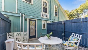 Un jardin avec une maison bleue, une table blanche, une chaise blanche et une poubelle blanche.