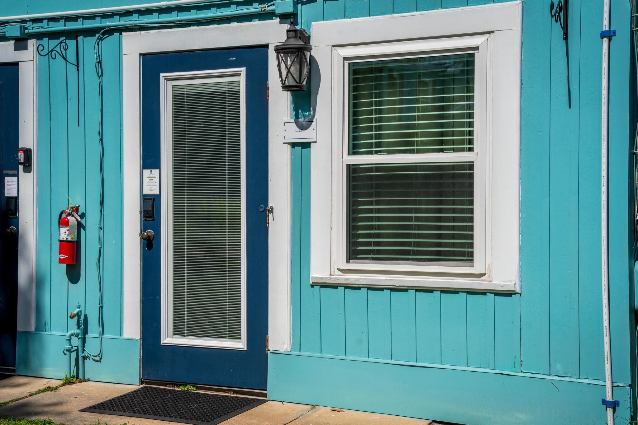 A teal building with a blue door, a window with blinds, a fire extinguisher, and a black lantern.
