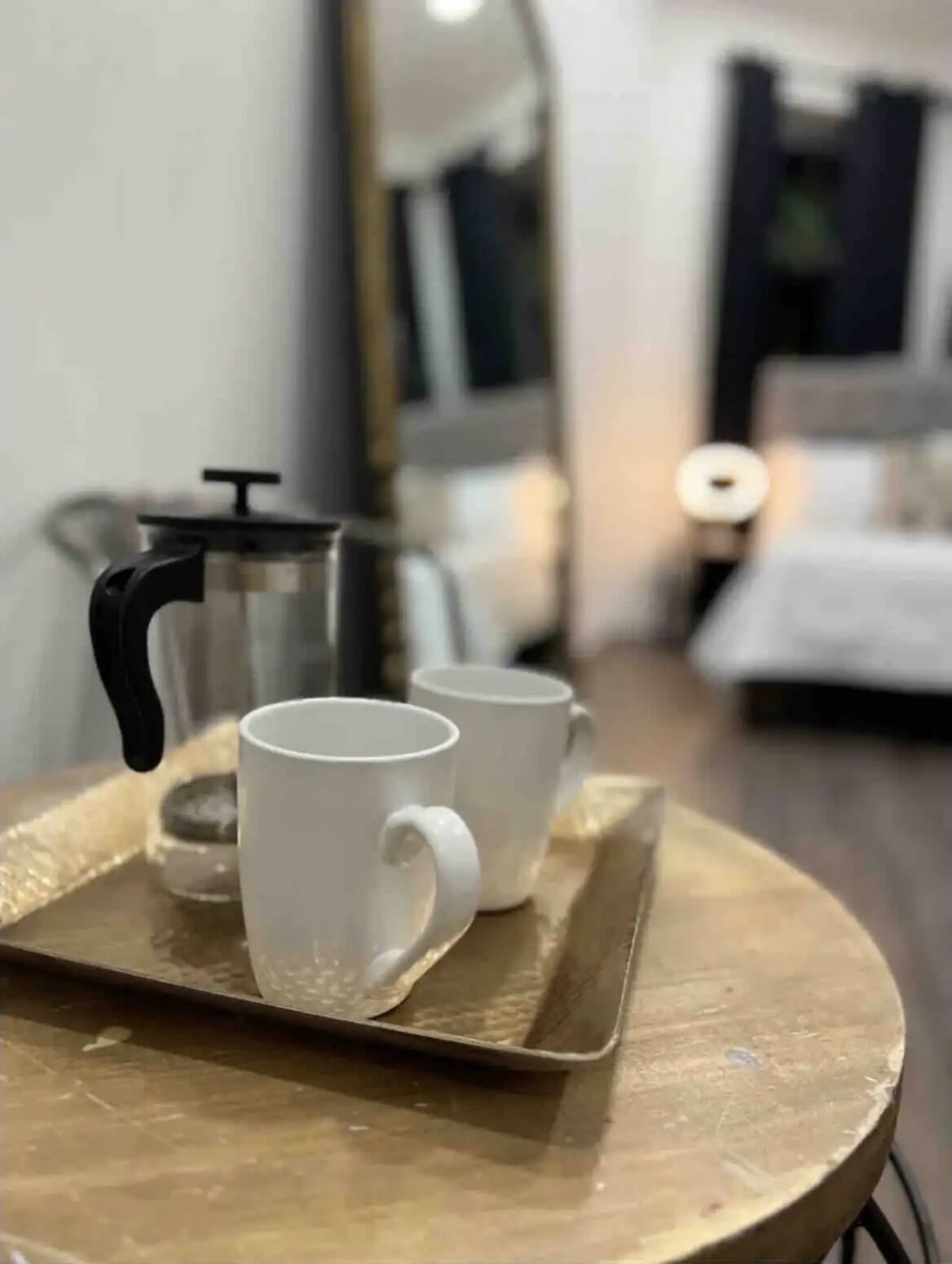 Un plateau avec deux tasses blanches posées sur une table ronde, une cafetière et un miroir qui reflète une pièce avec un lit et une table de chevet.
