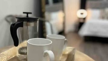 Un plateau avec deux tasses blanches posées sur une table ronde, une cafetière et un miroir qui reflète une pièce avec un lit et une table de chevet.
