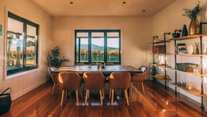 Un comedor con una mesa grande y sillas, vistas a viñedos a través de amplias ventanas, y una estantería con diversos objetos.