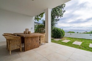 Enjoy casual outdoor dining on the patio with a rustic table and wicker chairs, surrounded by a well-manicured lawn and hedge.