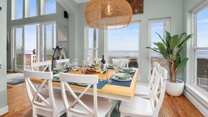 Uma sala de jantar com vista para o oceano, cadeiras brancas, uma mesa de madeira e um lustre.