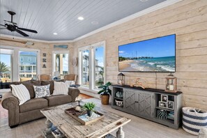 A living room with a large TV, wooden walls, a ceiling fan, and a view of the sea. - 2nd Row Beach 6BR | Sleeps 15 | Pointe West Amenities (Galveston)