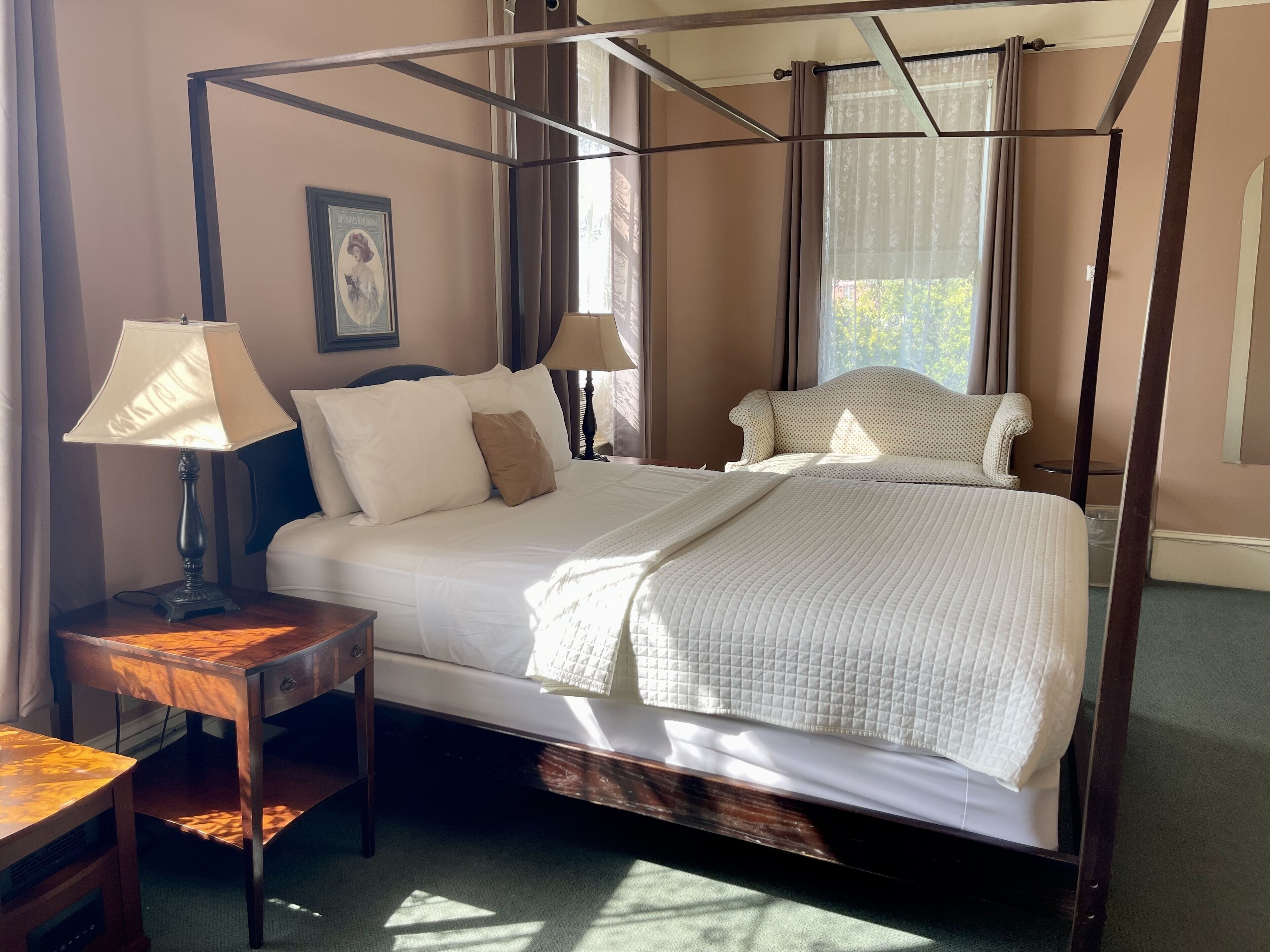 A four-poster bed with a canopy, two bedside tables, a framed picture on the wall, and a view of the outdoors through the windows.