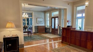 A hotel lobby with wooden flooring, a reception desk, a lamp, a vase with flowers, and a framed picture on the wall.