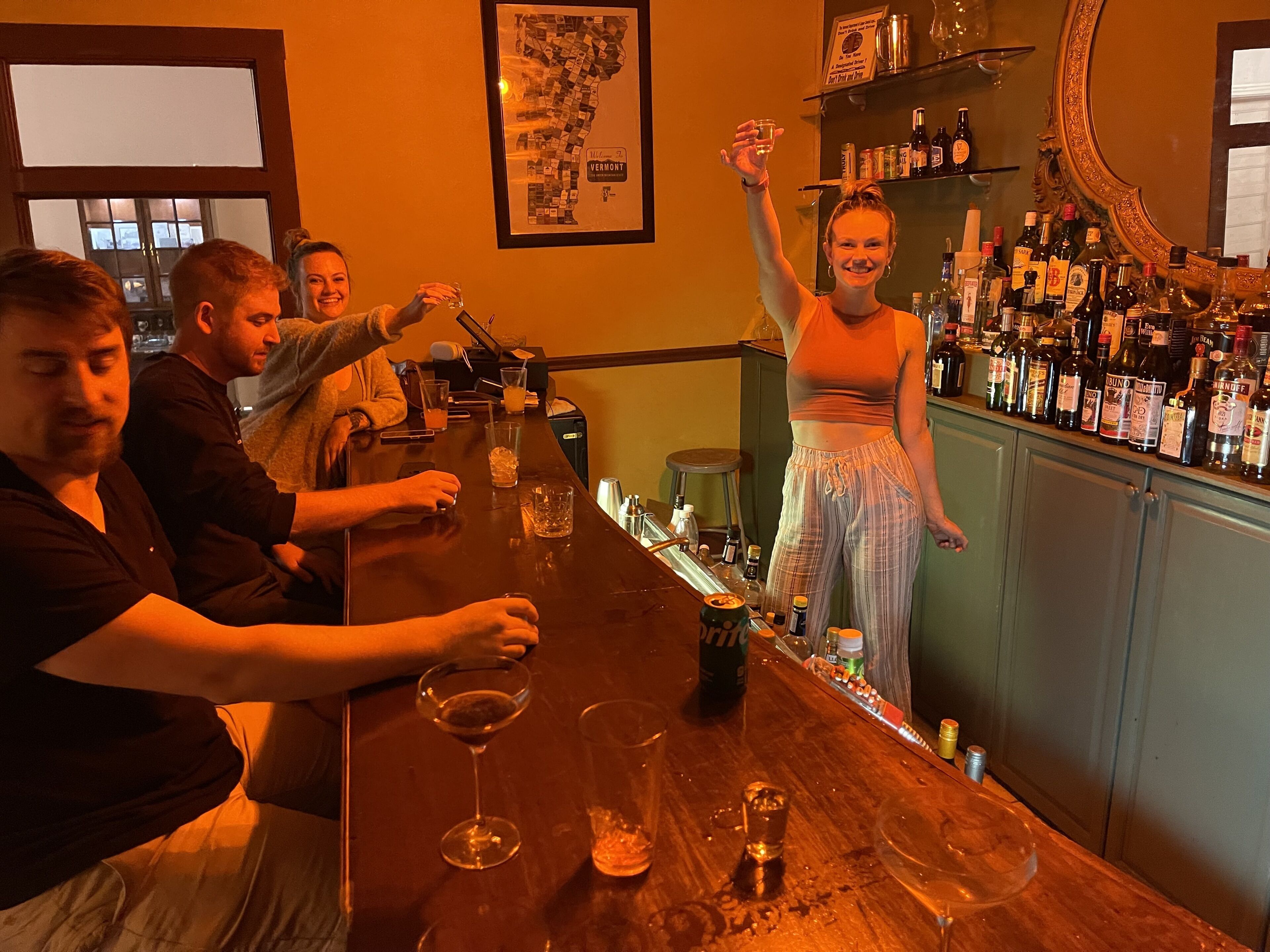 A bar area with a wooden counter, various bottles, glasses, and a map on the wall.