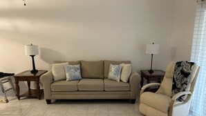 En stue med en beige sofa, to sofaborde med lamper, et mønstret tæppe på en stol og et flisegulv.
