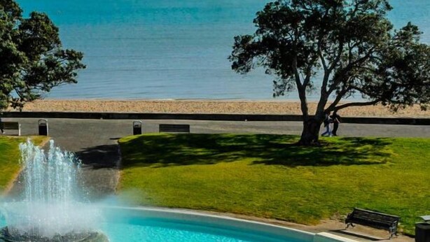 A fountain with two statues in a pool, surrounded by grass and trees, with a bench and a pathway nearby. - Mission Bay Beach House. Pet-Friendly (Auckland)